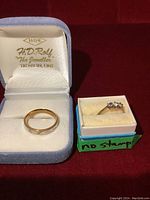 Two rings displayed in separate jewelry boxes; a flat band inscribed 'True Love' in a blue box and a three-stone ring marked 'no stamp' in a white box.