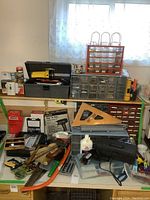 Overview photo showing several organizer bins with small plastic drawers, black plastic toolbox, various hand tools (hammers, saws), glue stick machine, bottles of liquids, and other assorted tools on a table.