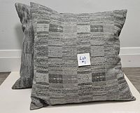 Two decorative pillows showing front and angled view. Gray geometric block pattern and feather-filled softness visible.