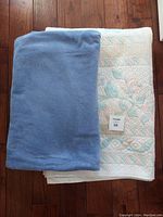Two folded blankets laid flat on wooden floor: one solid light blue fleece blanket folded on the left, and one quilted bed cover with pink and blue floral and geometric pattern underneath.
