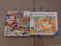 Two boxed jigsaw puzzles side by side on a dark surface: a Ravensburger 500-piece puzzle showing cats and a dog, and a 1,000-piece puzzle with a colorful city street scene.