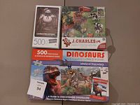 Photo showing three puzzle sets: Ghostbusters 500-piece, J. Charles 550-piece farm scene, and Dinosaurs educational set with puzzle and book.
