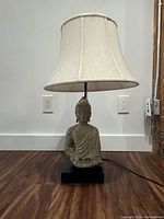 Full view of Buddha lamp showing Buddha statue base, fabric lampshade, black rectangular base, power cord.