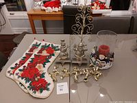 Christmas decorations including 2 poinsettia stockings, 2 silver stocking hooks with decorative figures, gold bell garland, silver metal ornament tree, and a large glass pillar candle holder with snowman themed base.