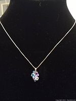 Necklace on black bust showing the fine silver chain and multi-colored gemstone cluster pendant with blue, purple, and clear stones.