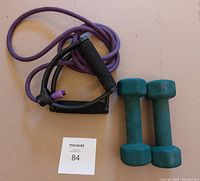 Photo showing a purple resistance band with black foam handles and two green dumbbells on a beige surface.