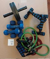 Top-down image showing 2 resistance bands (green and red with black handles), sets of blue dumbbells (3 lb and 5 lb), ab wheel with green wheel, and black push-up bars with blue foam grips all arranged on a flat surface.