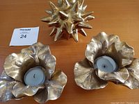 Two gold flower-shaped tea light holders with a white candle in each, and a gold decorative spike ball on a wooden surface.