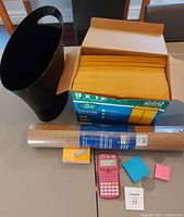 Photo of a black plastic trash can, manilla envelopes in a box, a rolled corkboard, pink calculator, sticky notes, and staples on a table