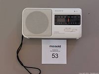 Front view of Sony portable radio showing speaker grille, tuning dial with AM/FM bands, and wrist strap attached