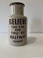 Close-up photo of the beige ceramic jar showing the detailed embossed inspirational text on its side.