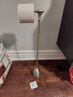 Photo shows a single weighted toilet roll holder with a toilet paper roll on it, placed on hardwood floor against a gray wall. There are packages visible on the sides in the background.
