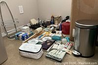 Photo showing assorted toiletries, creams, lotions, and small items spread on floor next to a metal bath waste bin and shower head rack.