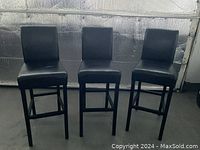 Three black Parsons bar stools arranged side by side against a metallic background, showing bonded leather upholstery with some visible wear.