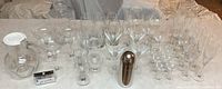 Photo showing an assortment of clear glassware and drinkware including martini, margarita, wine, champagne, and water glasses, along with two clear glass pitchers and a stainless steel martini shaker arranged on a cloth-covered surface.