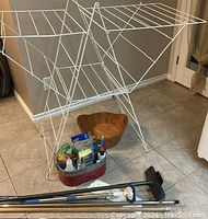 Full view of white metal laundry drying rack, red metal tub with cleaning supplies, wooden bowl, and clothes hangers on floor.