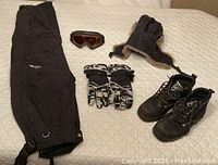 Photo showing all items: black North Face ski pants, Kombi ski gloves, Oakley ski goggles, Roots black winter boots, and brown winter hat with ear flaps.