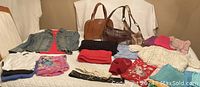 Photo showing a variety of women's clothing including sweaters, tops, pants, and three handbags arranged on a surface.