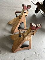 Pair of rusty steel car jackstands with triangular bases and sawtooth support tops, showing wear and rust, placed on a concrete floor.