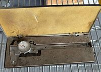 Dial caliper stored inside a worn case with fabric or foam lining, showing signs of writing and surface wear.