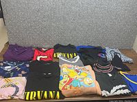 Various men's, women's, and youth tops laid flat showing graphics and colors including Nirvana and The Simpsons graphics.