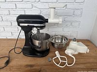 Full front view of the KitchenAid stand mixer with two bowls, meat grinder attachment, paddle, hook, and whisk accessories shown on wooden table against white brick wall.