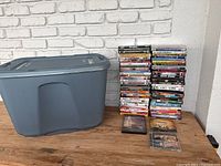 Blue plastic storage tote with lid next to tall stacks of DVDs on wood surface against white brick background.