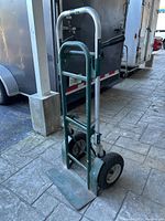 Green two wheel dolly showing frame, handle, and foot plate, positioned upright on pavement.