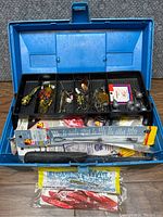 Open blue Umco tackle box showing multi-compartment plastic tray with various fishing lures, hooks, weights, and fishing line, plus lower compartment with tools.