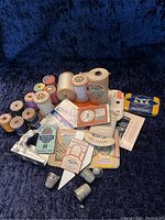 Overview of variety of antique sewing materials including multiple thread spools, needle packets, and thimbles arranged on dark blue fabric.