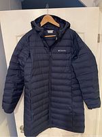 Full front view of the navy blue Columbia ladies hooded down coat hanging on a door, showing the full length, hood, and side zip pockets.