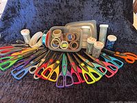 Wide angle photo of all 25 scissors spread out with rolls of ribbons and punch-out shapes in containers behind them