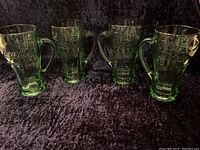 Four green tinted glass mugs with embossed Coca Cola logo, arranged in a row against black fabric.