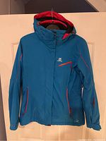 Front view of blue Salomon ski jacket with hood, showing red zipper accents and chest pocket.