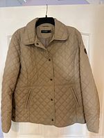 Front view of beige quilted jacket hanging on black hanger showing the diamond pattern and snap button closure.