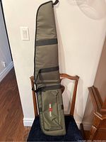 Full view of the green and black padded scoped gun case standing upright on a chair. Shows the entire case length and handle.