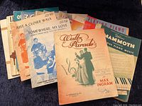 Spread of multiple vintage piano music books and sheet music, showing different titles and illustrations.