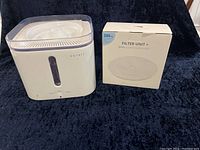 Petkit Eversweet pet drinking fountain next to box of two replacement filters on dark fabric background.