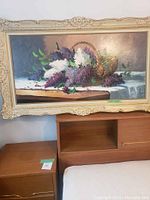 Frontal image of a floral still life painting featuring lilacs and basket on a white tablecloth, framed ornately in cream.