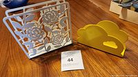 Two decorative napkin holders on a wooden surface, one white metal with floral design and one yellow cloud-shaped holder.