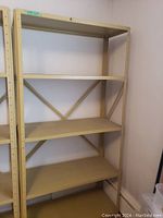 Full view of beige metal shelving unit with four wooden shelves and triangular metal cross braces at back, placed against a white wall.