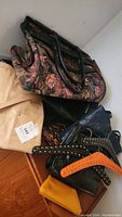 Photo showing various bags piled together on a floor including floral bag, beige bag, blue bag, yellow pouch, and the studded leather hangers.