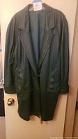 Full front view of vintage green leather jacket hanging on door, showing length, lapel collar, and front pockets.