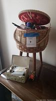 Photo of the vintage sewing basket with lid open showing multiple spools of thread inside. The basket is fabric lined with a red padded lid and woven body, shown on wooden tripod stand.