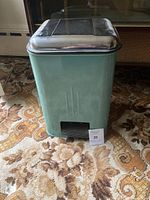 Front view of vintage turquoise metal garbage can on patterned carpet, showing foot pedal and body details.