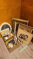 Box of assorted vintage picture frames, some empty, some containing old photographs or prints, placed on floor against wood walls.
