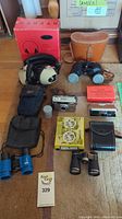 Full view of lot showing vintage camera, multiple binoculars, headphones, film rolls, and cases on carpeted floor