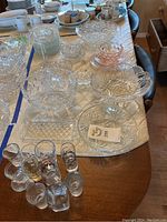 View of entire lot including multiple glass and crystal bowls, plates, and various shot glasses arranged on a table with blue tape sections