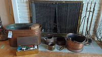 Full view of firewood holder, small copper bucket, brass kettle, metal ash container, and box of matches with other fireplace items on hearth in front of screened fireplace.