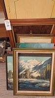 Photo showing a stacked group of framed paintings including a snowy mountain landscape and other artworks